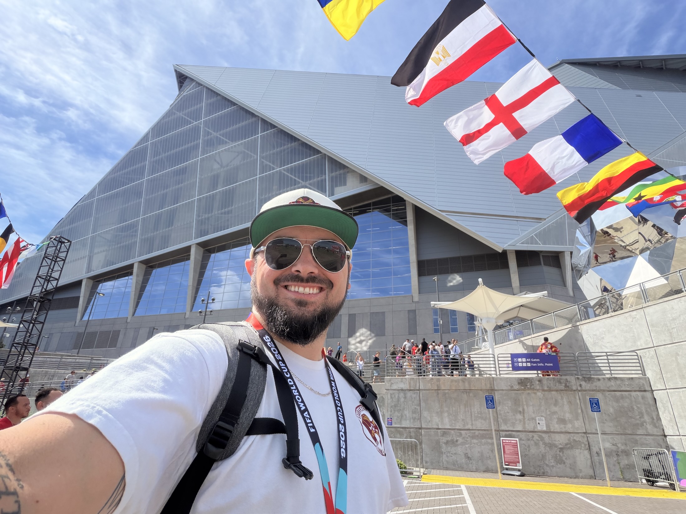 Tyler Pilgrim in sunglasses and a FIFA World Cup 2026 lanyard takes a selfie outside Mercedes-Benz Stadium in Atlanta, with national flags including England, France, and Egypt strung overhead.